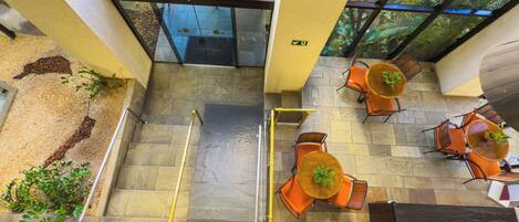 An upper-level view of a hotel lobby with a glass ceiling, outdoor seating area, and a staircase.