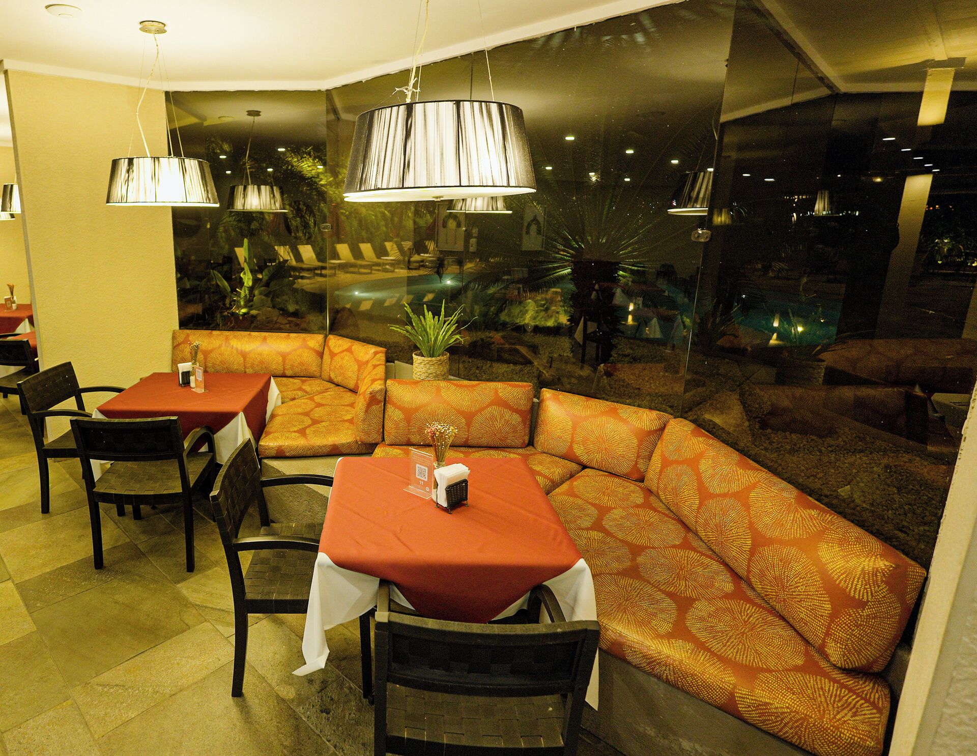 A restaurant with a corner booth seating area, tables with red tablecloths, and hanging pendant lights.