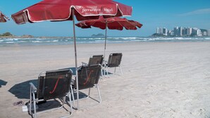 Ein Strand mit roten Sonnenschirmen, auf denen 'Ferrarretto Hotel' steht, Metallstühlen und einer Stadtkulisse im Hintergrund.