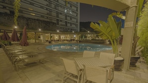 Ein Poolbereich mit Liegestühlen und Sonnenschirmen, umgeben von einem mehrstöckigen Gebäude und einem Restaurant.