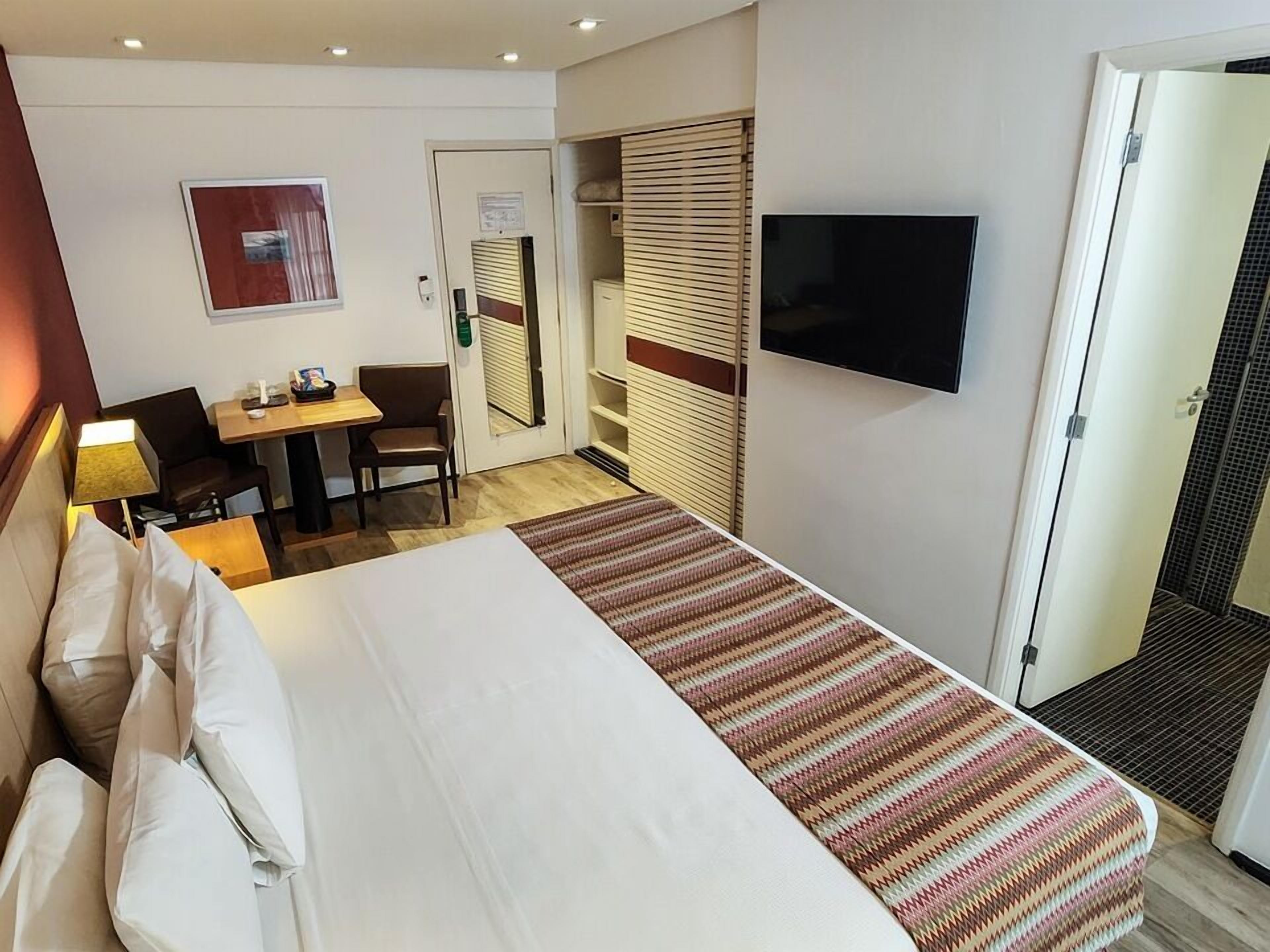 Una habitación de hotel con cama, escritorio con silla, televisor montado en la pared y puerta que da a un baño.