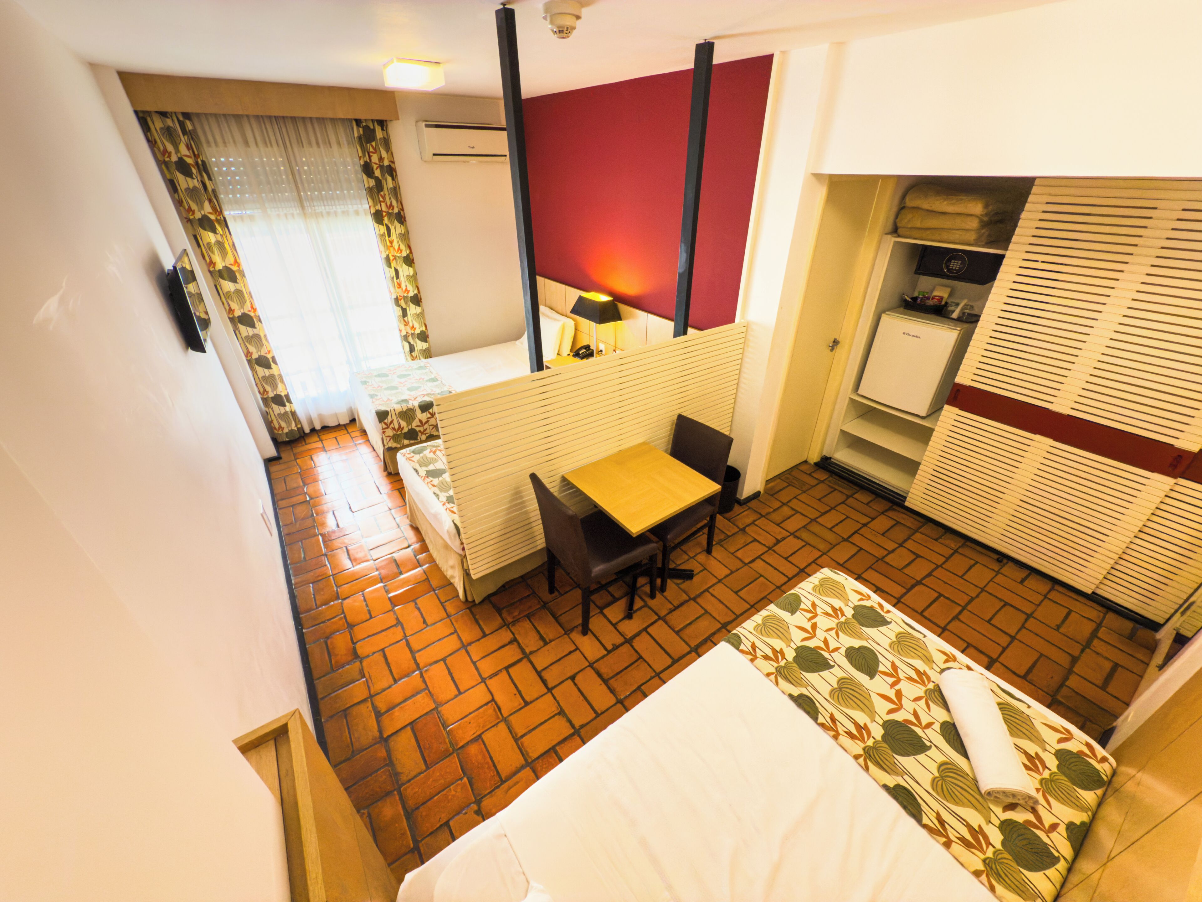 A hotel room with a bed, a desk, a chair, a small table, a wardrobe, and a bathroom with a toilet and sink.