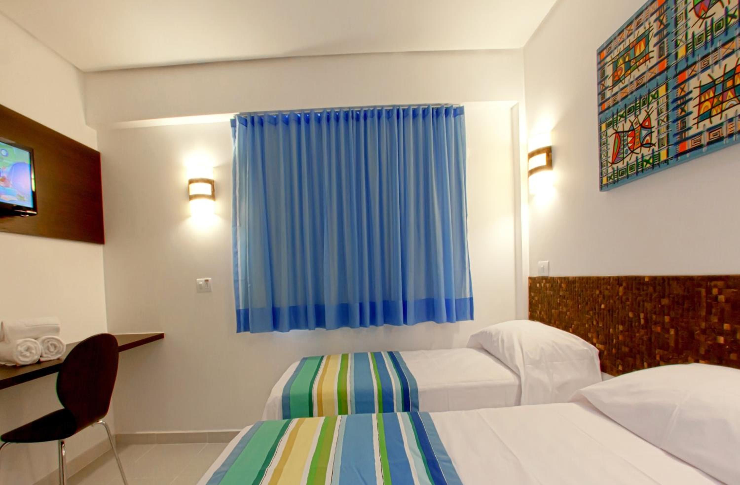 A hotel room with two beds, a desk with a chair, a television, and a large window with blue curtains.