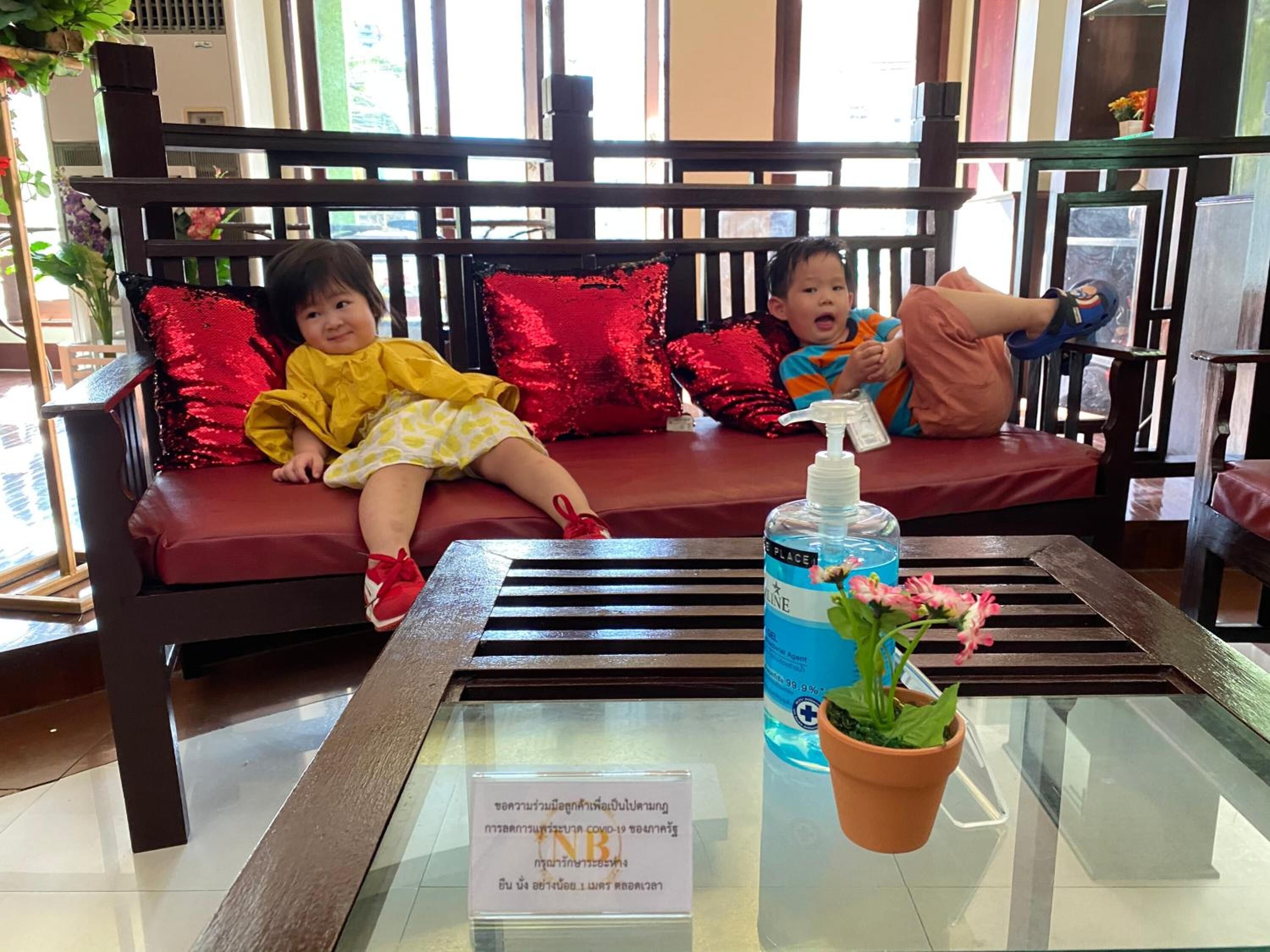 A seating area with a glass table in front, a bottle of hand sanitizer, and a sign with Thai text.