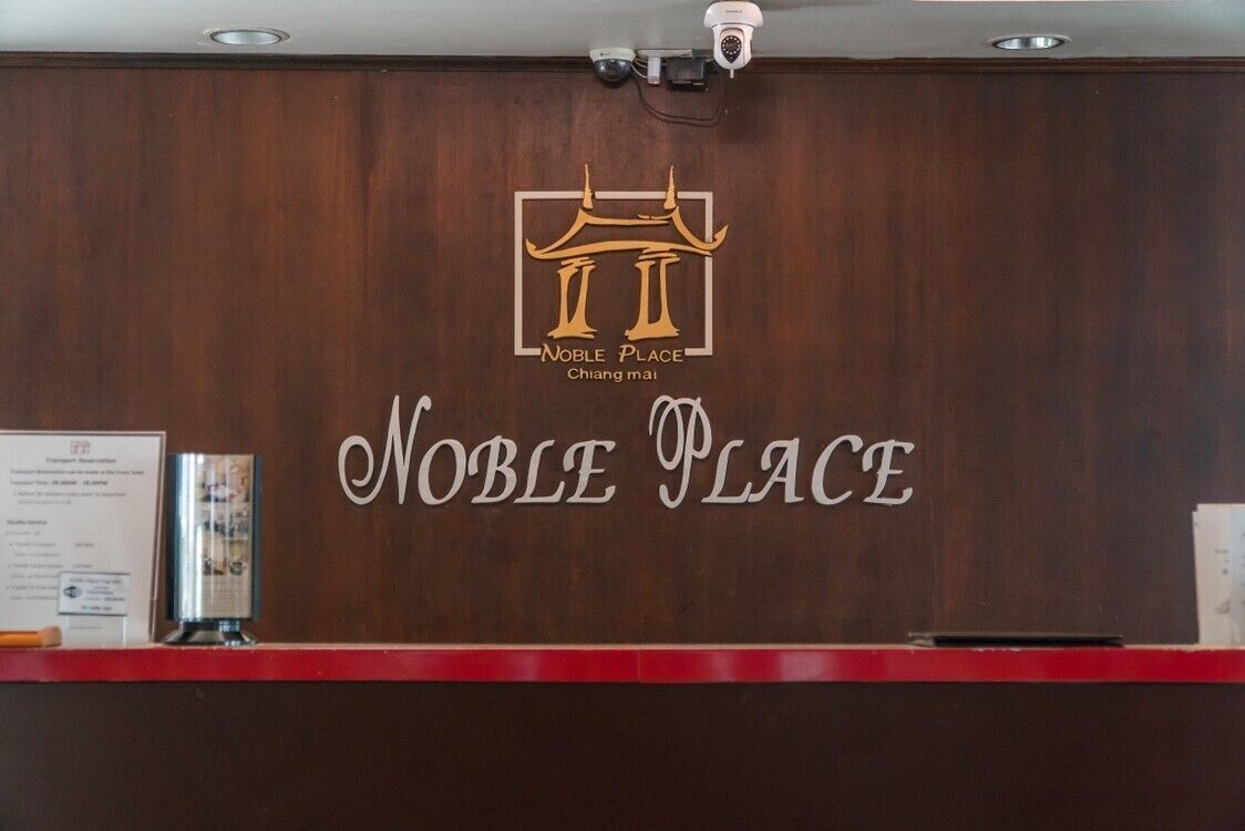 A reception desk with a wooden panel background, a logo with 'Noble Place' and 'Chiang mai' text, and a security camera above.
