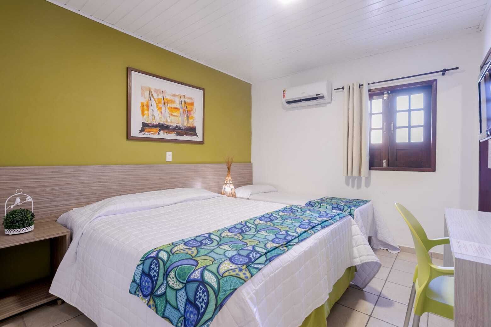 A hotel room with a bed, a desk, a chair, a window with curtains, an air conditioning unit, and a painting on the wall.