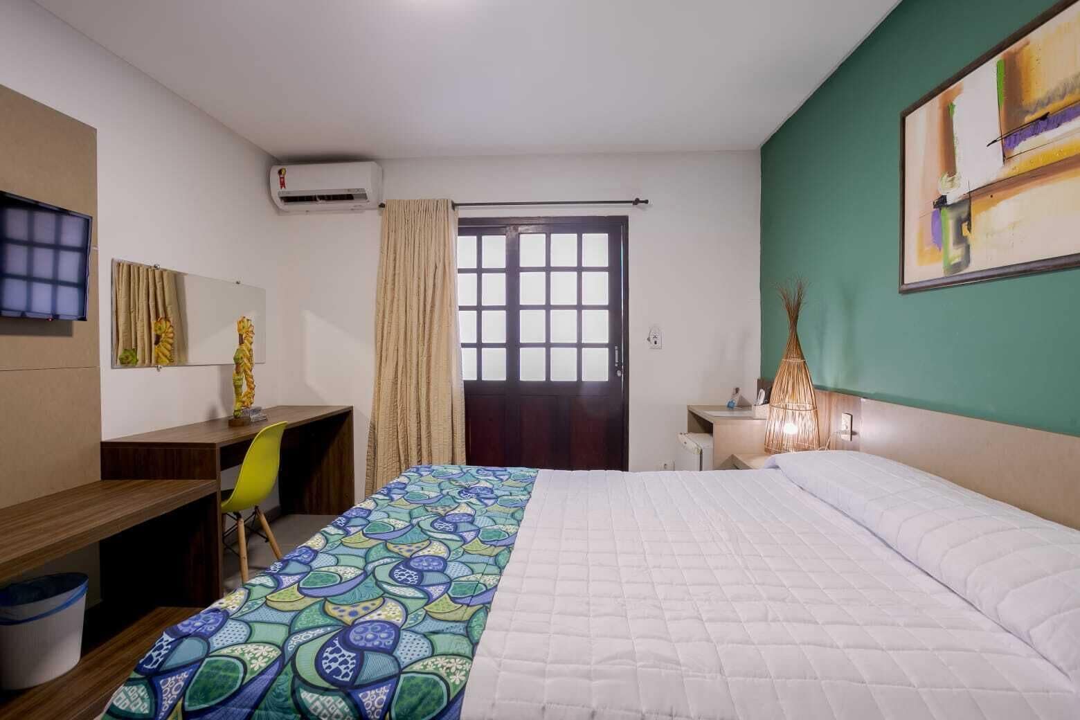A hotel room with a bed, a desk, a chair, a TV, an air conditioner, a door, and a painting on the wall.