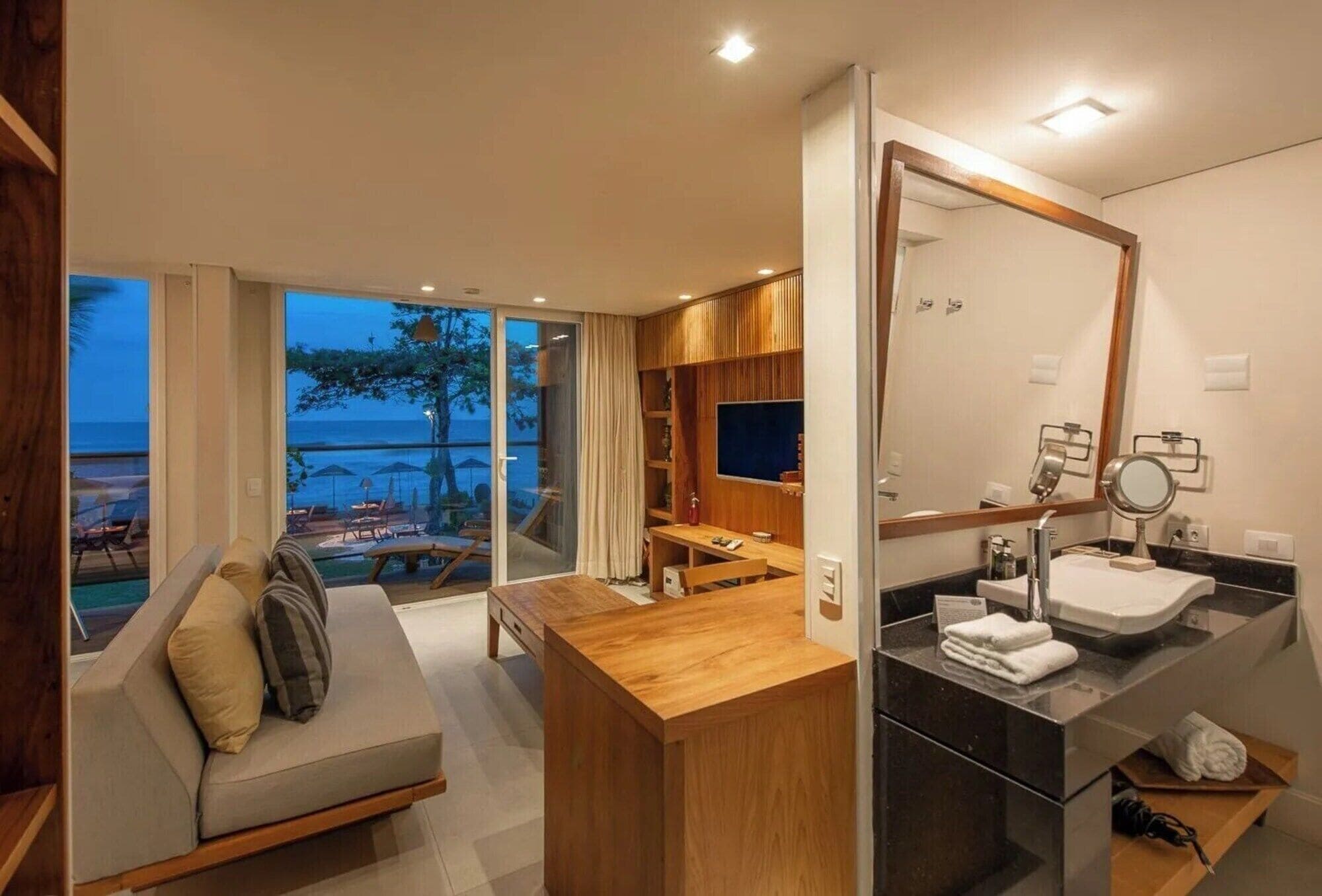 Quarto de hotel moderno com uma grande janela com vista para uma piscina e praia, uma secretária de madeira, um sofá, um banheiro com um espelho grande e uma pia.