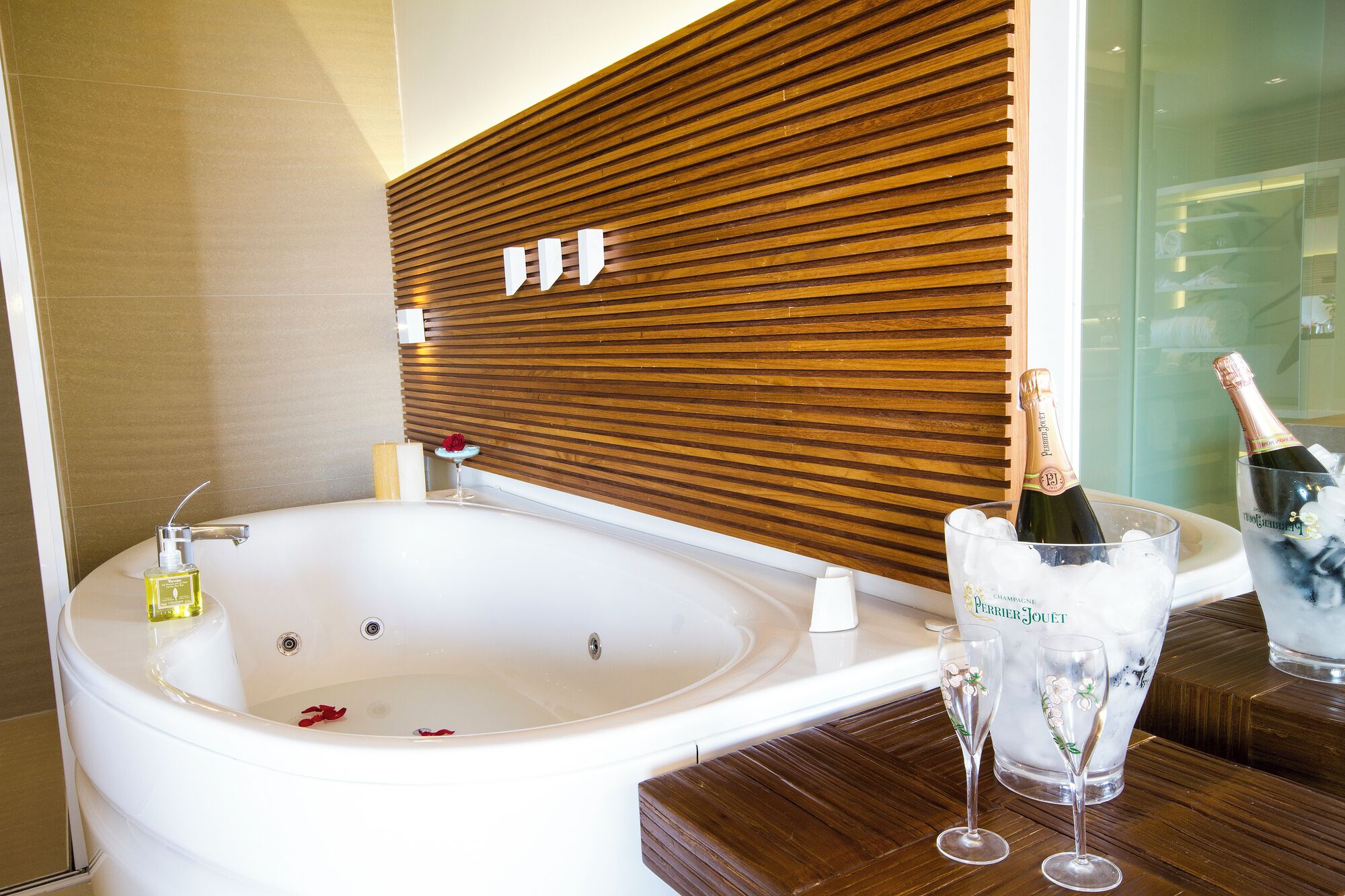A modern bathroom with a large bathtub, a wooden slatted wall, and a minibar with champagne and glasses.
