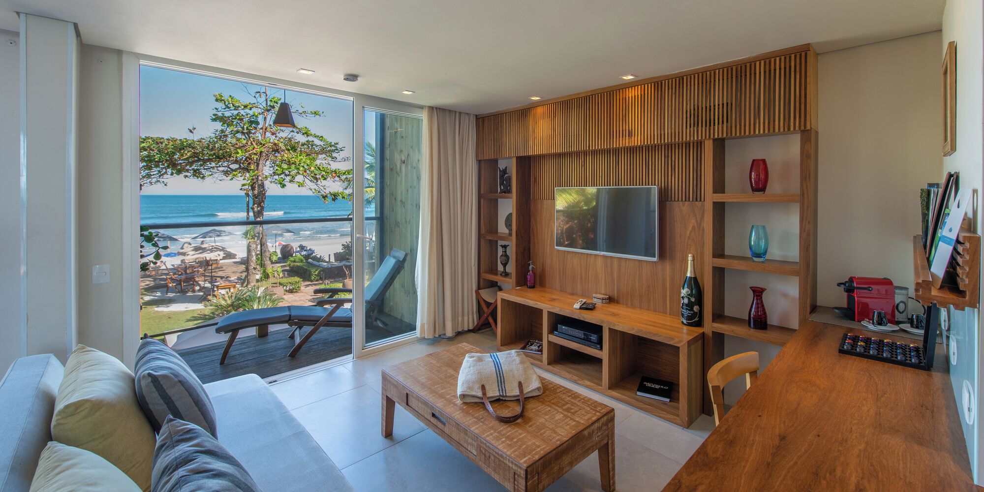 A modern living room with a large TV, wooden furniture, a coffee machine, and a view of the beach.