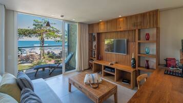 A modern living room with a large TV, wooden furniture, a coffee machine, and a view of the beach.