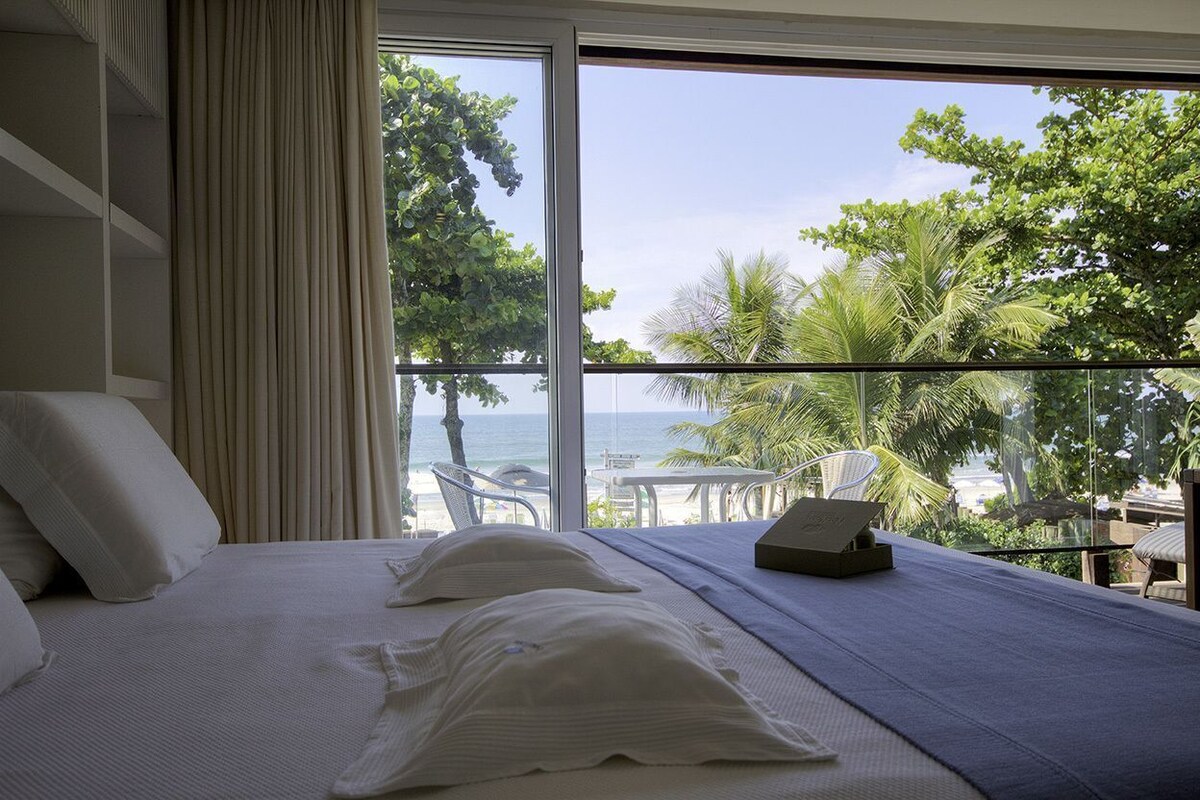A hotel room with a bed, a view of the ocean, and a balcony with chairs.