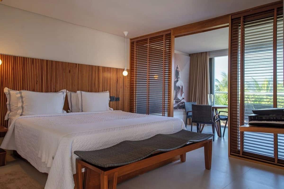 A spacious bedroom with a large bed, a wooden bench, and a view of the outdoors through a sliding door.