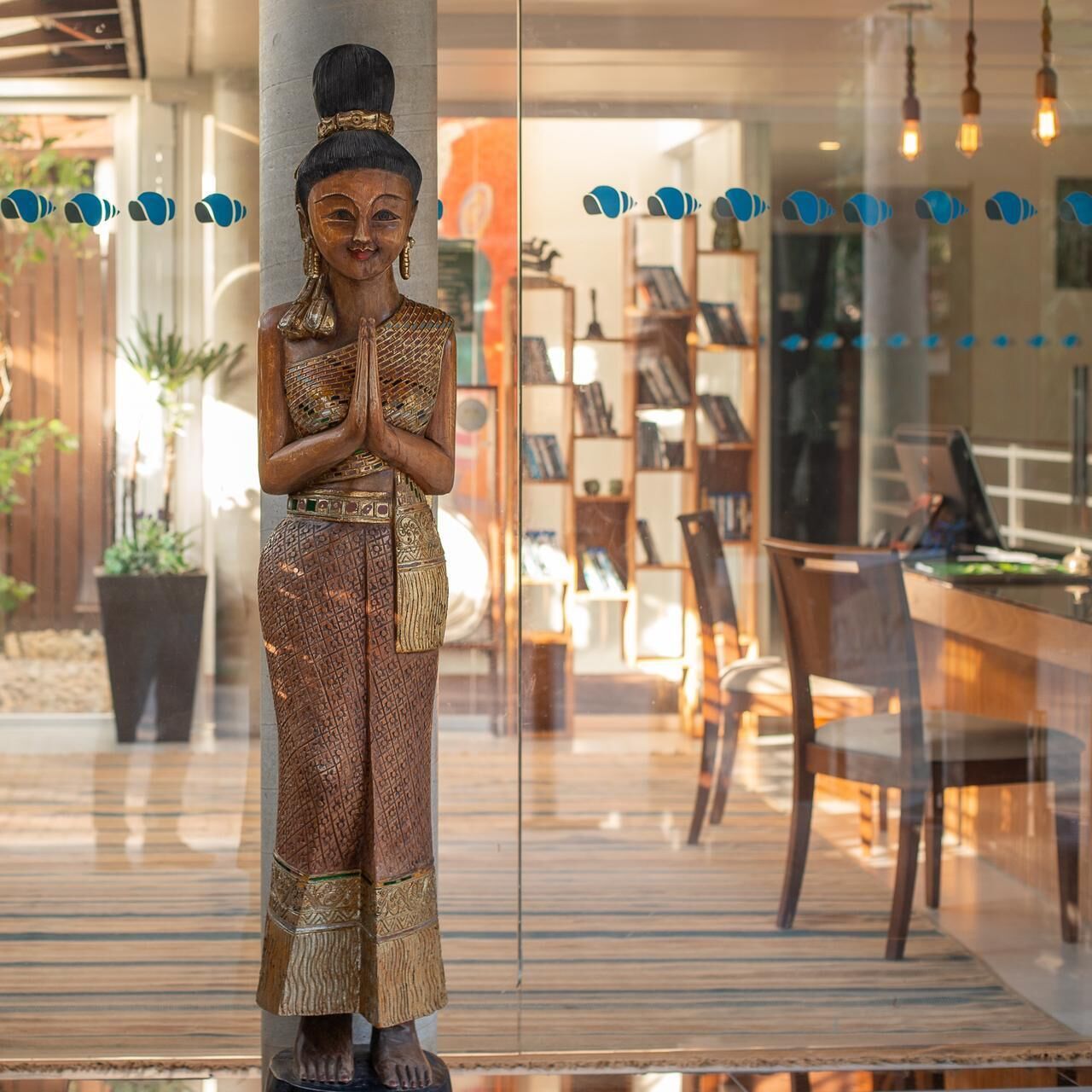 A statue of a figure in traditional attire, standing in a modern indoor setting with a glass partition, wooden furniture, and a visible outdoor area with plants.
