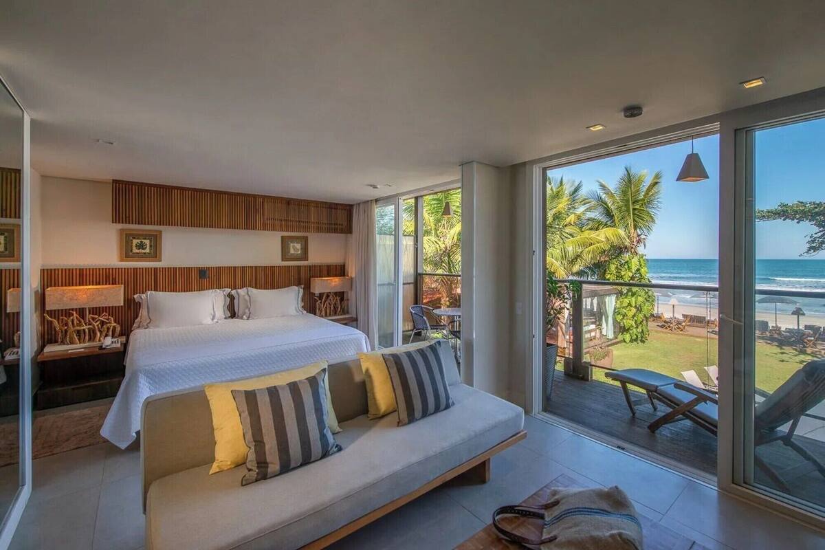 A modern bedroom with a large bed, a sofa, and a balcony with a view of the beach and ocean.