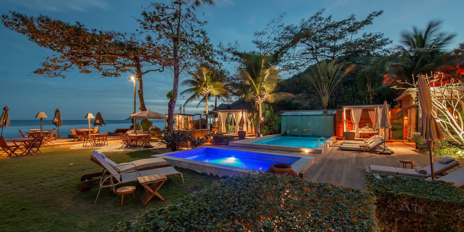 Zona de piscina con sillones de descanso y sombrillas, rodeada de plantas tropicales y vistas al mar.