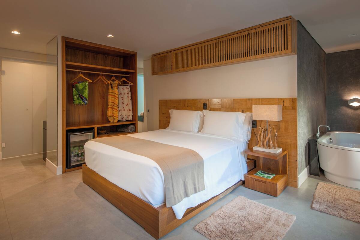 A modern bedroom with a large bed, a wooden wardrobe, a built-in desk, a bedside table, a nightstand, a small refrigerator, and a bathtub.