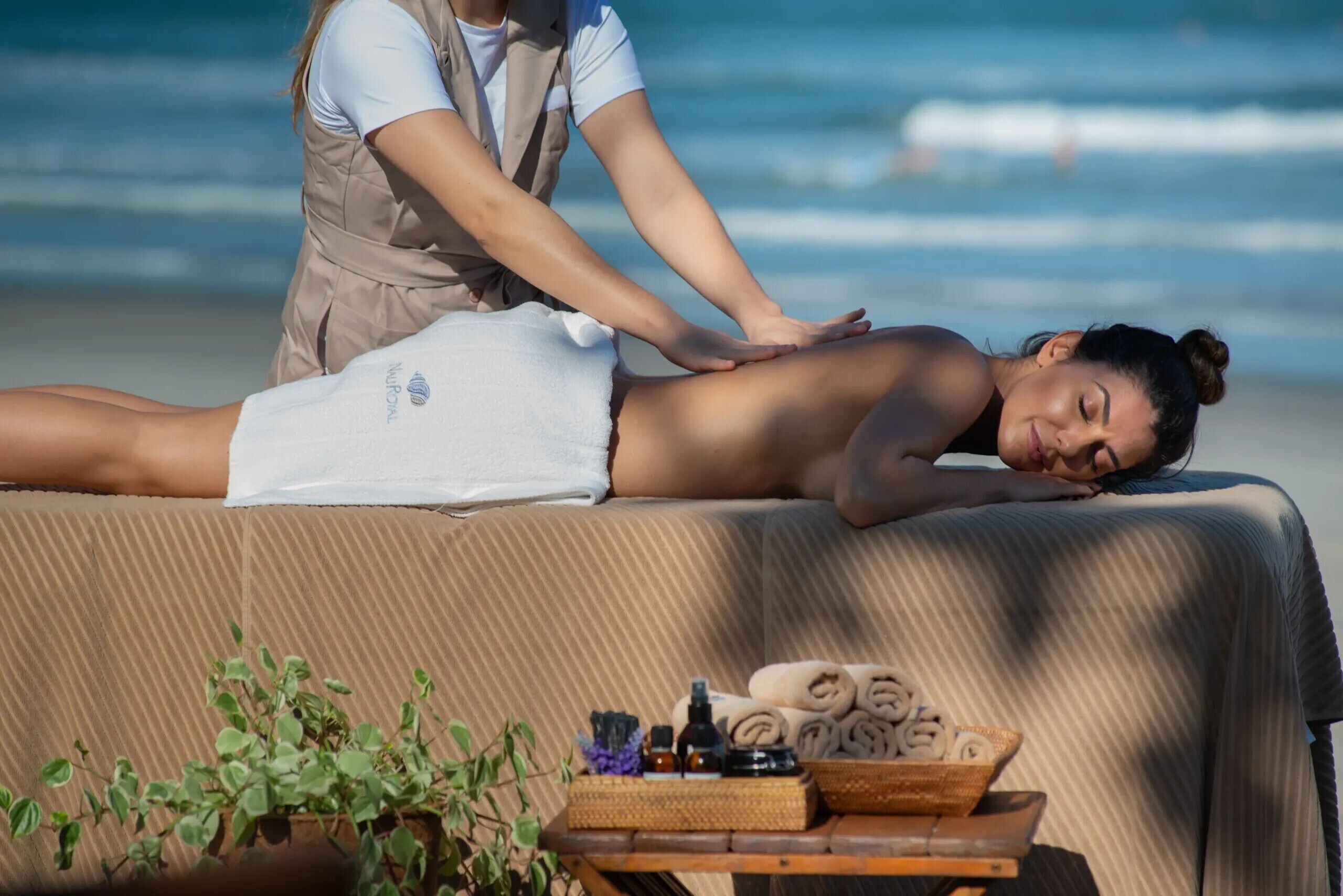 A massage therapy session on a beach with a towel-covered massage table, a small table with towels and bottles, and a potted plant.