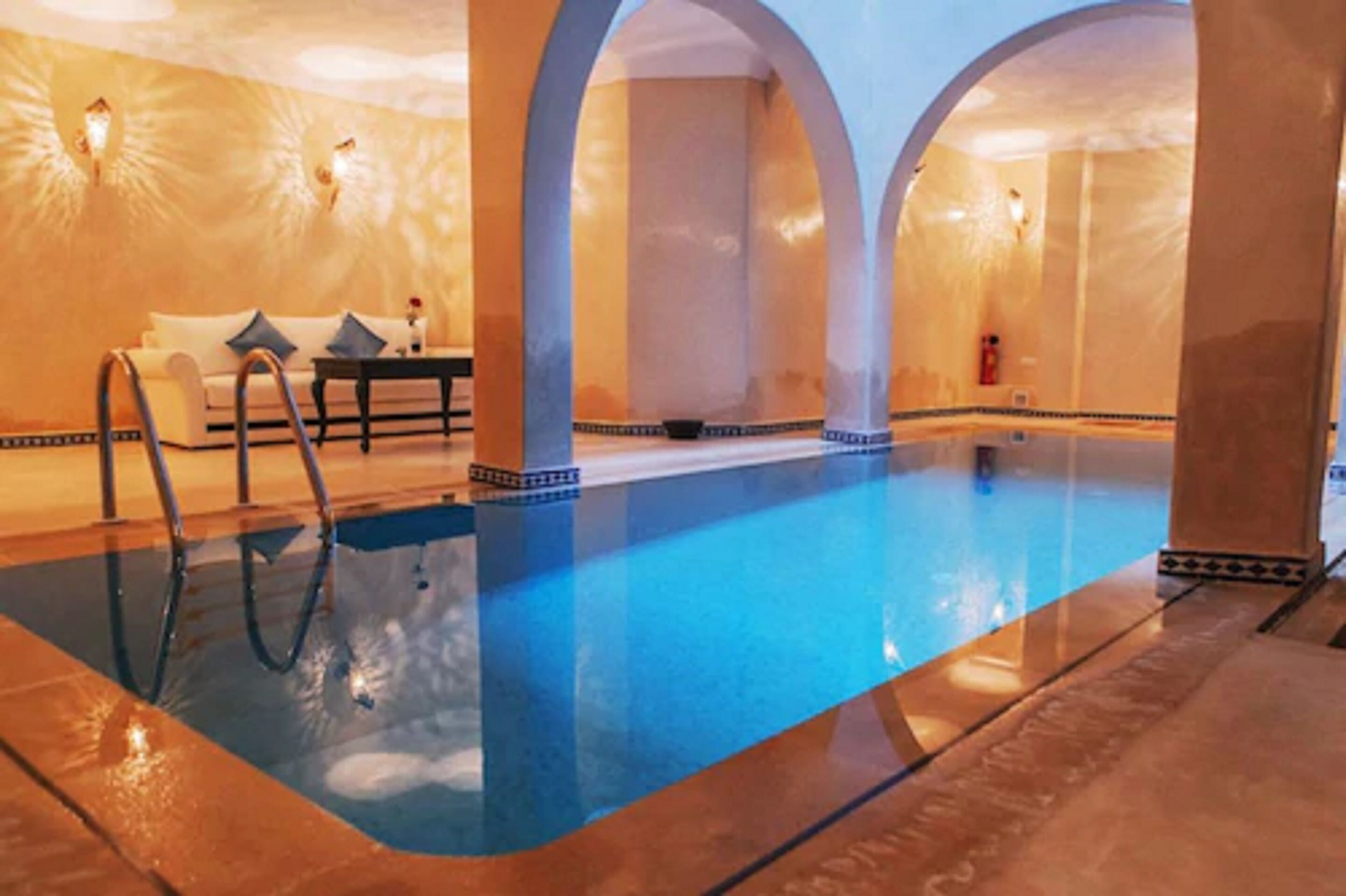 Une piscine intérieure à l’eau d’un bleu cristallin, entourée de colonnes blanches et agrémentée d’un canapé blanc avec des coussins.