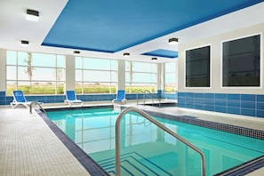 En indendørs swimmingpool med krystalklart, blåt vand, omgivet af blå og hvide flisevægge, og store vinduer med udsigt til det fri.