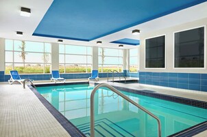 Una piscina interior con agua azul cristalina, rodeada de paredes de azulejos blancos y azules, y amplias ventanas que ofrecen vistas al exterior.