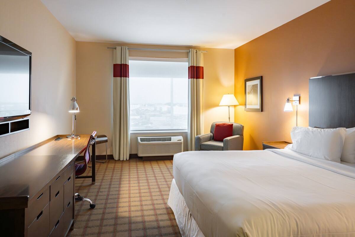 A hotel room with a large bed, a desk with a chair, a flat-screen TV, a window with curtains, and a city view.