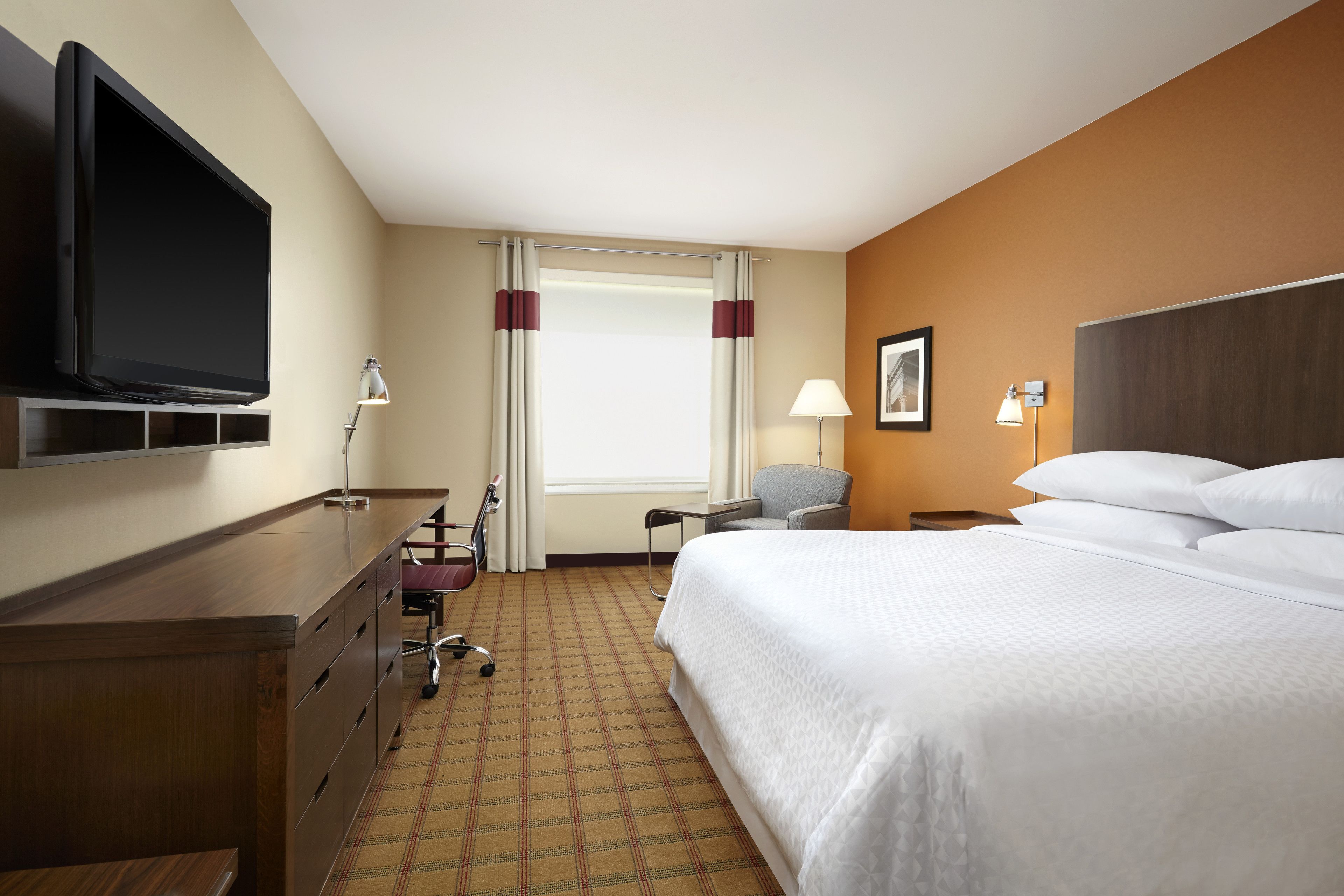Ein Hotelzimmer mit einem Bett, einem Schreibtisch mit Flachbildfernseher, einem Sessel, einer Schreibtischlampe und einem Fenster mit Vorhängen.