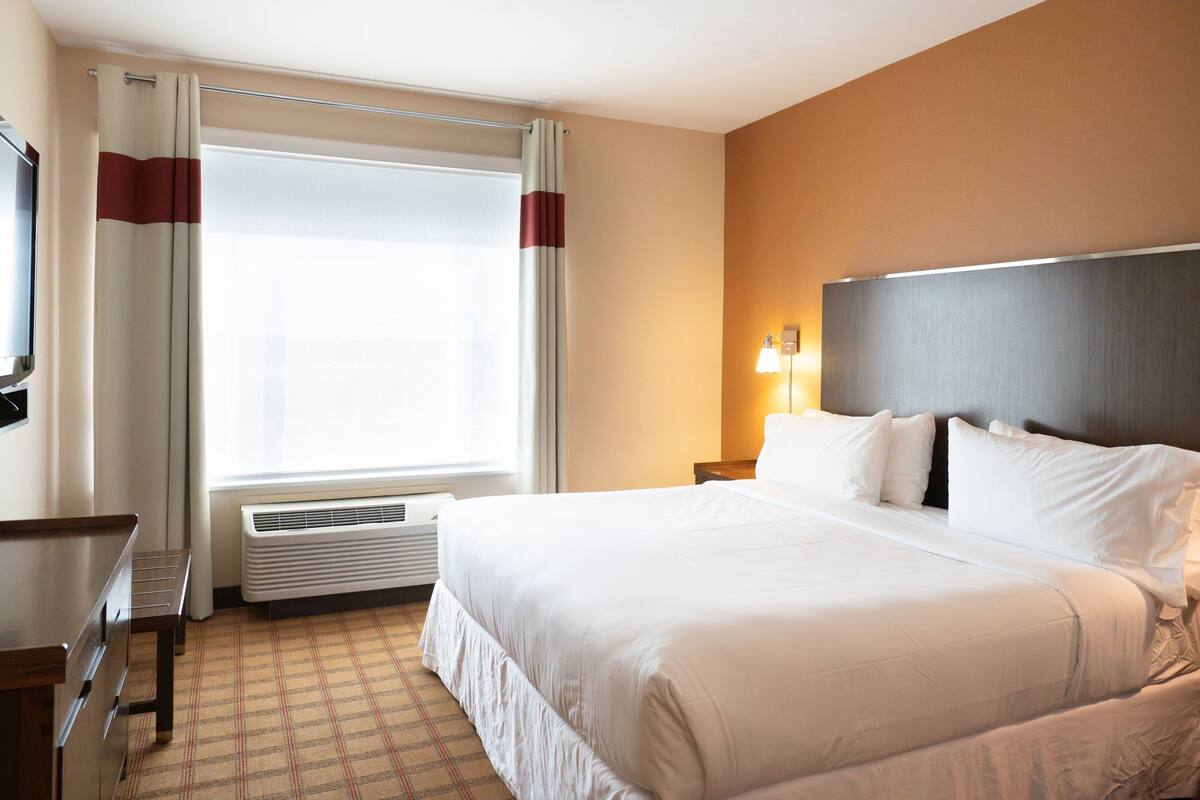 A hotel room with a large bed, a desk, a chair, a window with curtains, a wall-mounted TV, and a bedside lamp.