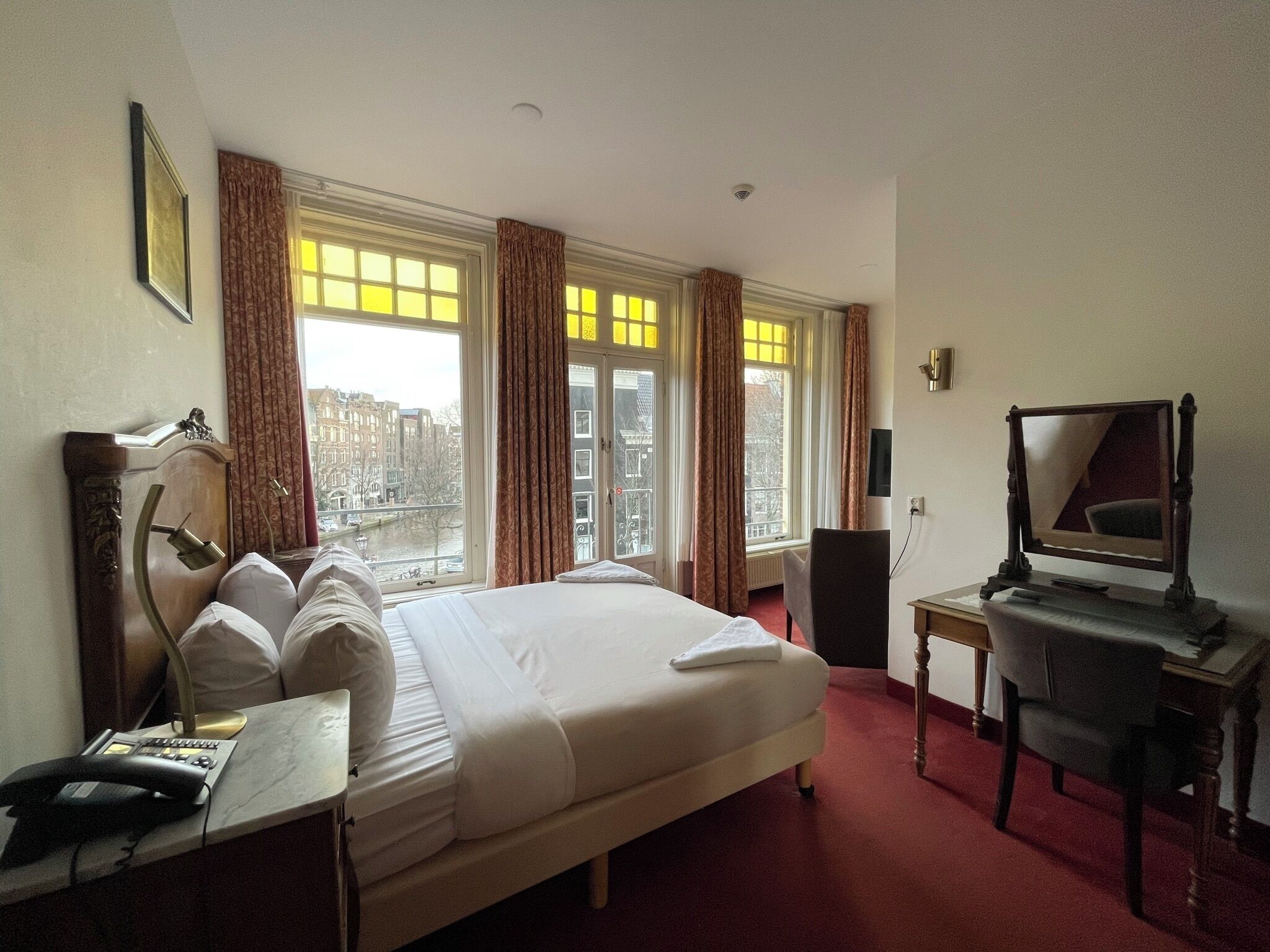 Habitación de hotel con una cama grande, un escritorio con teléfono, una silla, una lámpara de escritorio, un espejo y vistas de la ciudad a través de amplias ventanas.