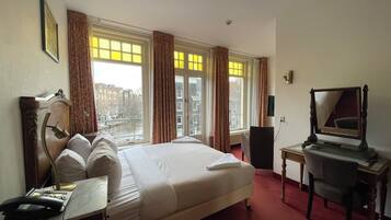 Habitación de hotel con una cama grande, un escritorio con teléfono, una silla, una lámpara de escritorio, un espejo y vistas de la ciudad a través de amplias ventanas.