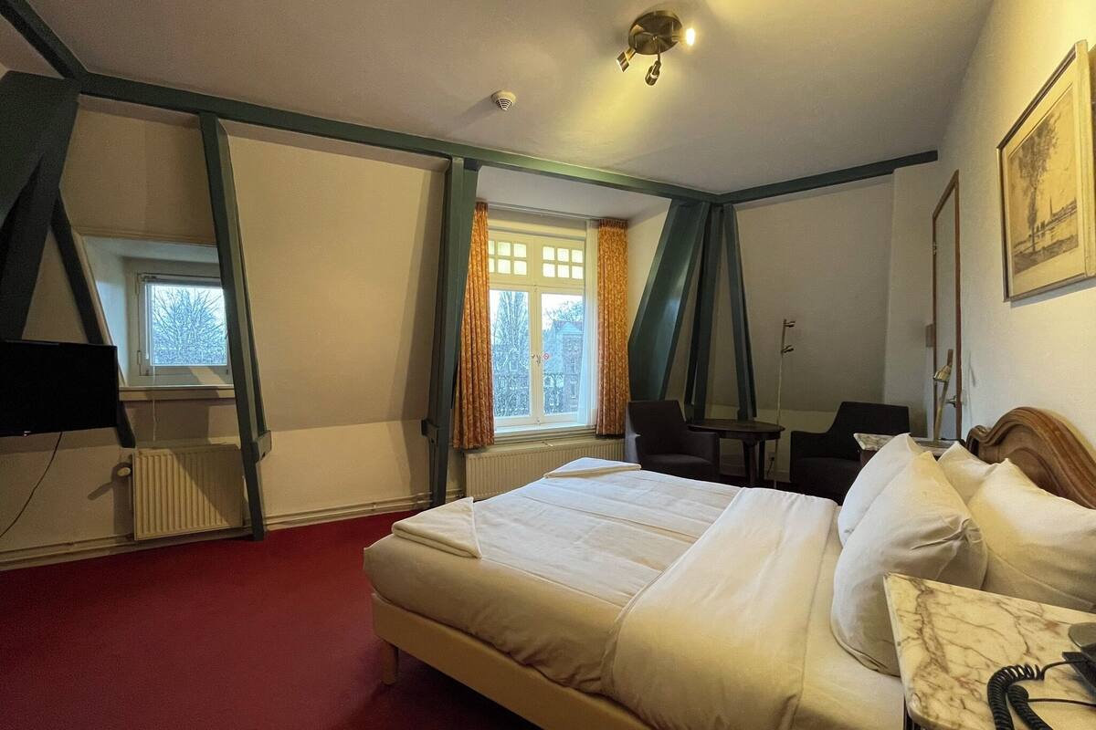 Ein Hotelzimmer mit einem Bett, einem Schreibtisch, einem Stuhl, einem Heizkörper, einem Fenster mit Vorhängen und einer Deckenleuchte.