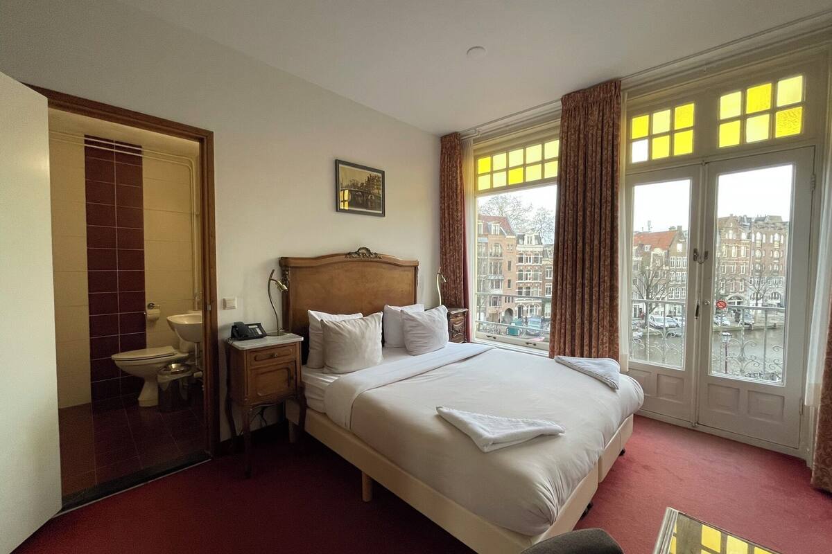 Ein Hotelzimmer mit einem großen Bett, einem Nachttisch mit Lampe, einem Fenster mit Vorhängen und Blick auf Gebäude.
