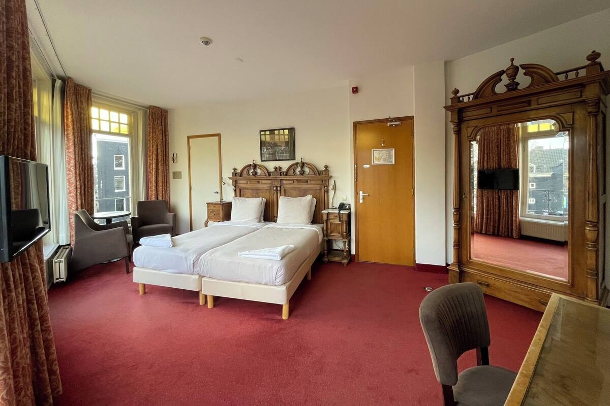 Ein Hotelzimmer mit einem Doppelbett, einem Schreibtisch mit Stuhl, einem Fernseher, einem großen Spiegel und einem Fenster mit Vorhängen.