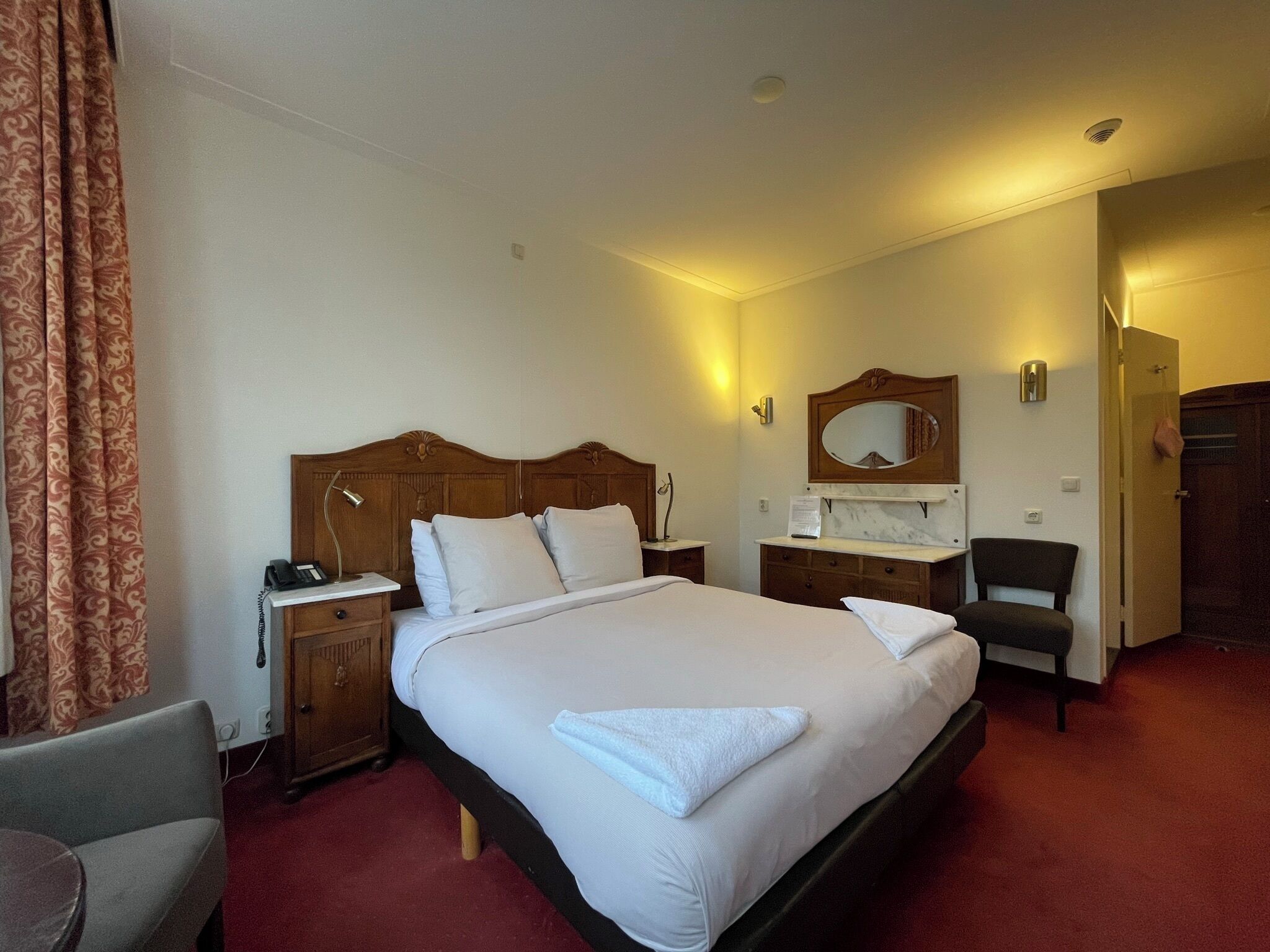 Habitación de hotel con una cama grande, mesitas de noche, una silla, un escritorio, un espejo y una puerta.