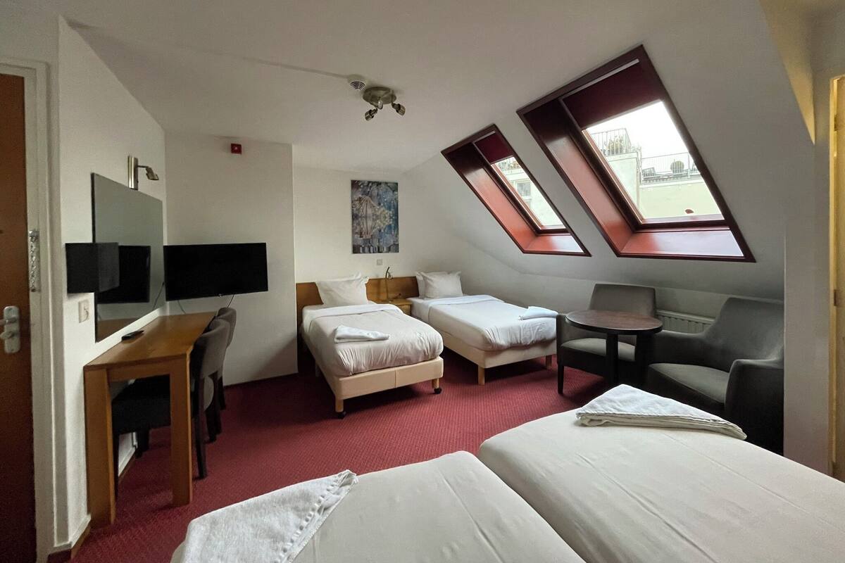 Ein Hotelzimmer mit zwei Betten, einem Schreibtisch mit Computer, einem Stuhl und einem kleinen Tisch. Dachfenster sorgen für natürliches Licht.