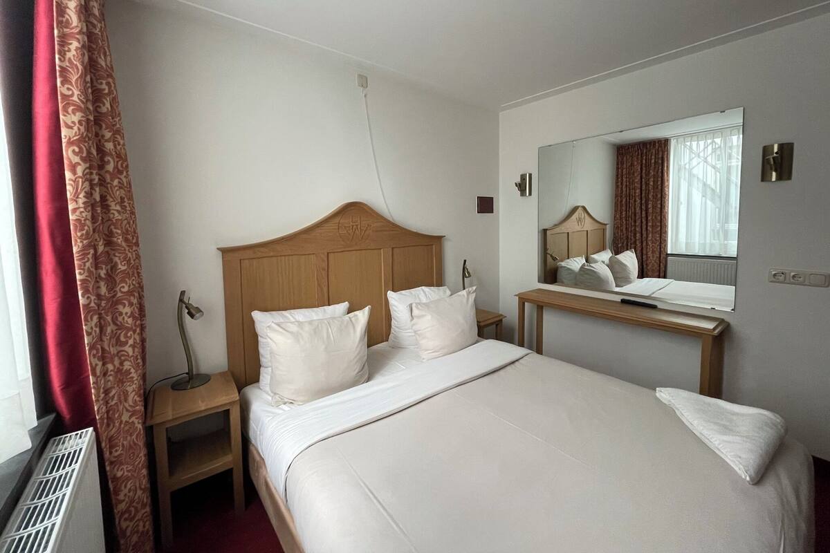 Ein Hotelzimmer mit einem hölzernen Kopfende, einem großen Spiegel, einem Nachttisch und einem Fenster mit Vorhängen.