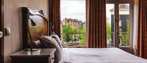 A hotel room with a large bed, a wooden nightstand, and a view of a cityscape through a window with curtains.