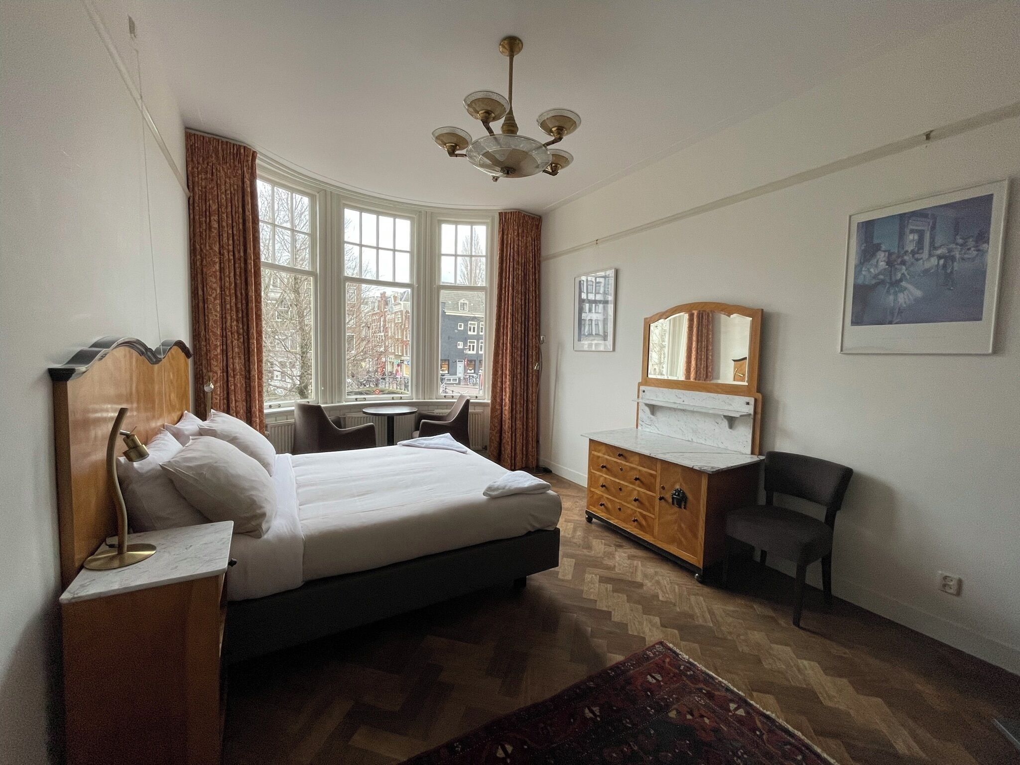 A hotel room with a bed, a dresser, a chair, a table, and a large window with a view of buildings.
