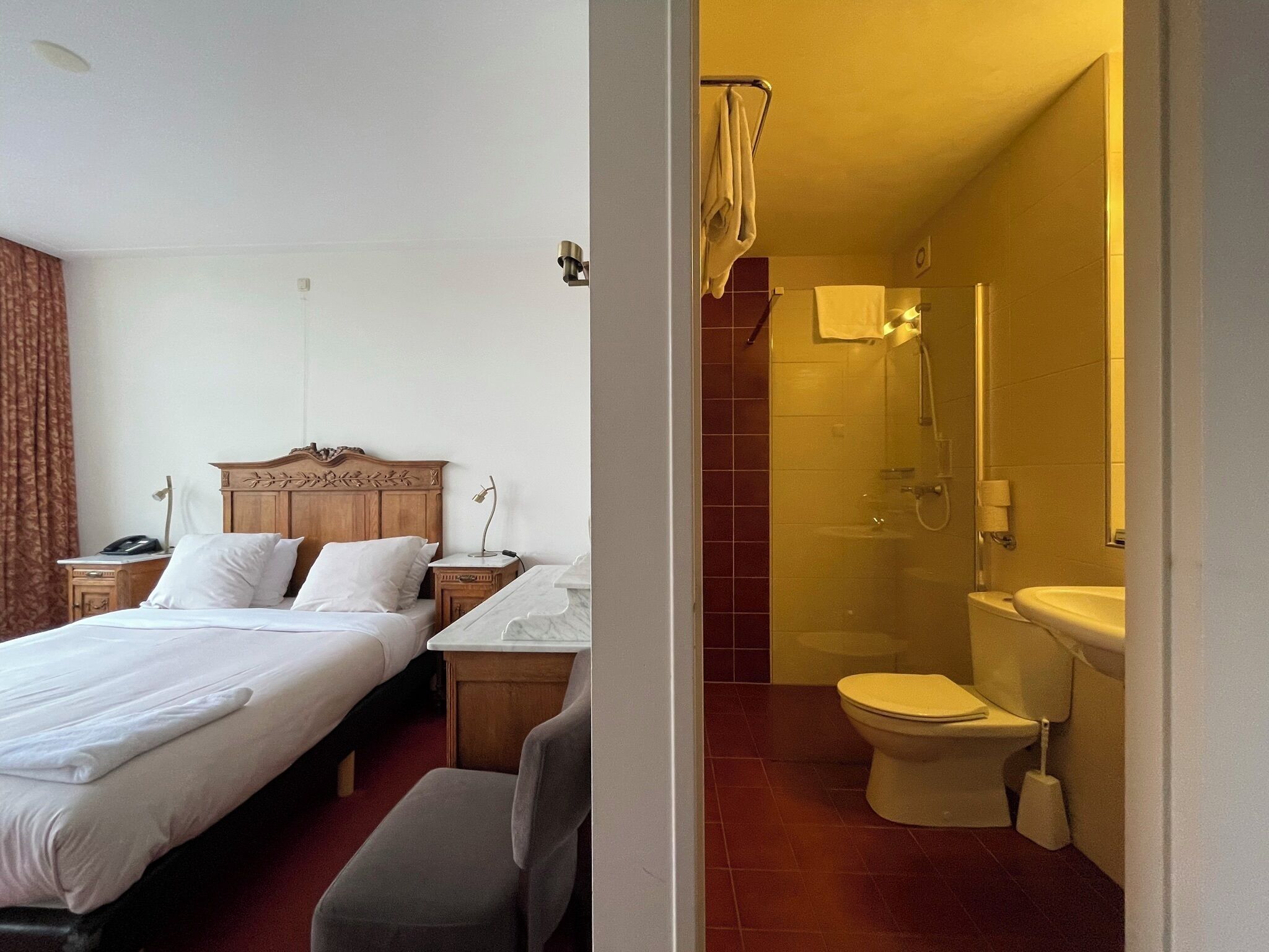 Habitación de hotel con una cama de madera, una mesita auxiliar de mármol, ducha y retrete.