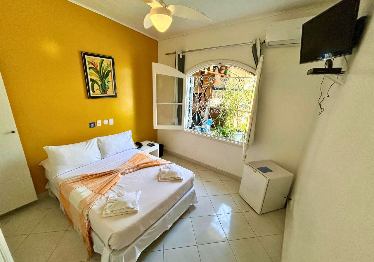 A hotel room with a bed, a bedside table, a TV, a window with a view, and a painting on the wall.