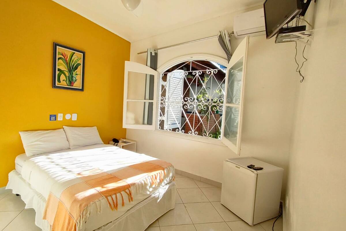 Une chambre à coucher avec un lit, une fenêtre ornée de barreaux en métal décoratifs, une petite armoire blanche et un téléviseur fixé au mur.