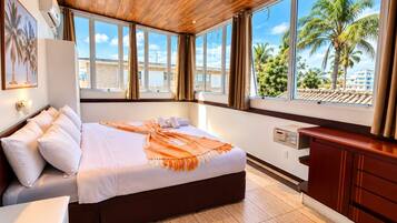 A hotel room with a large bed, a wooden ceiling, a view of palm trees, and a window with curtains.