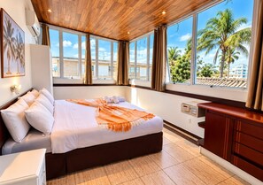 Ein Hotelzimmer mit einem großen Bett, einer Holzbalkendecke, Blick auf Palmen und einem mit Vorhängen drapierten Fenster.