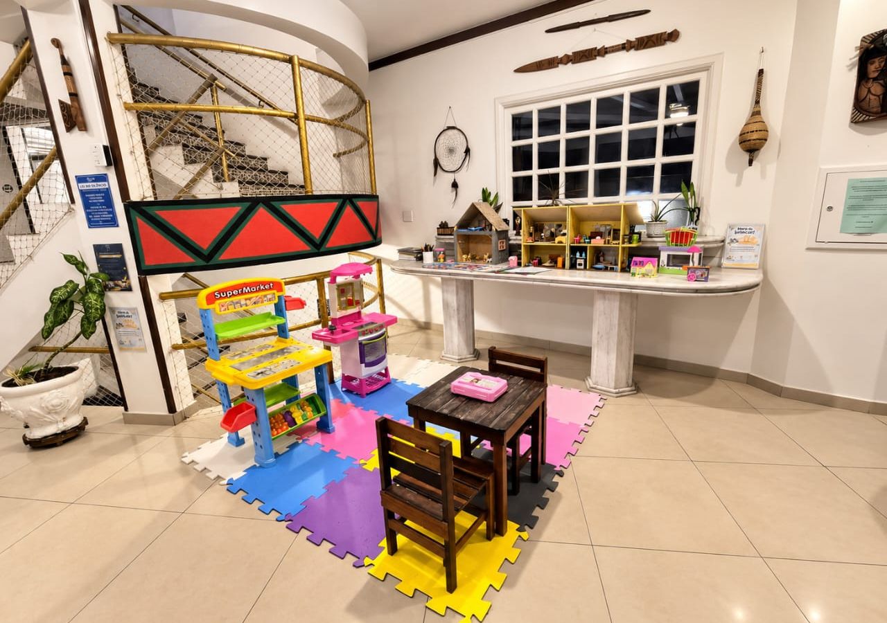 Espaço infantil com cozinha de brincar, loja de brinquedos e área de estar com mesa e cadeiras.