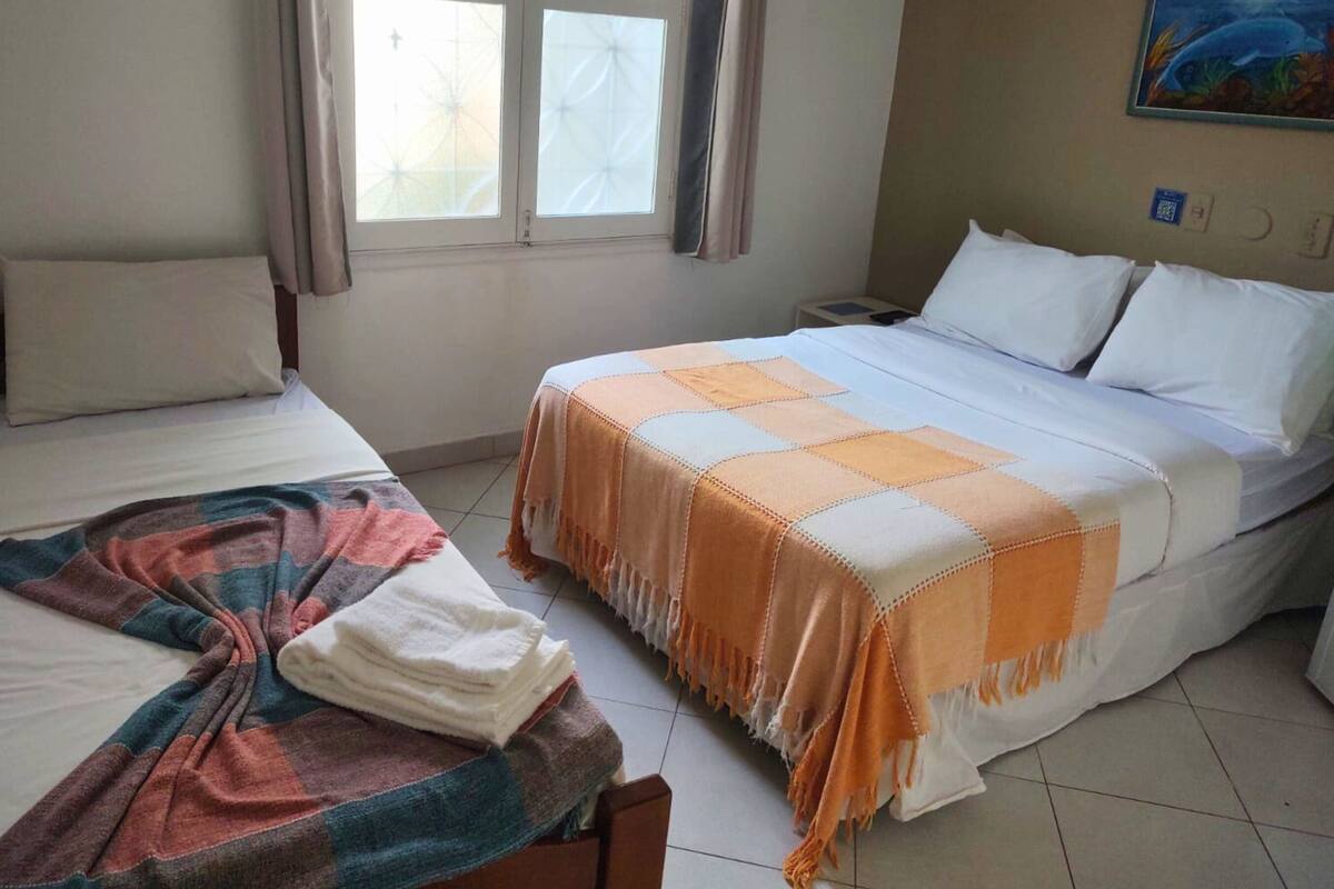Une chambre d’hôtel avec deux lits, une fenêtre avec des rideaux, un tableau au mur et un sol carrelé.