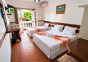 Quarto de hotel com duas camas, televisão, uma mesa pequena e varanda com vista para o verde.