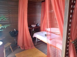 Quarto com piso de madeira, uma cama com lençol branco, uma mesinha, uma pia e uma planta.