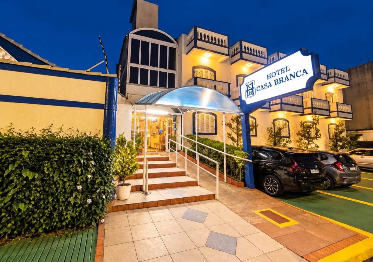 A hotel entrance with a sign reading 'HOTEL CASA BRANCA,' a covered staircase, and a parking area with cars.