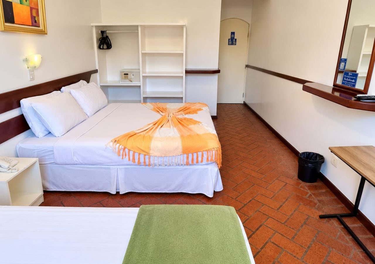 A hotel room with a bed, a bedside table, a green towel, a wooden bench, and a trash bin.