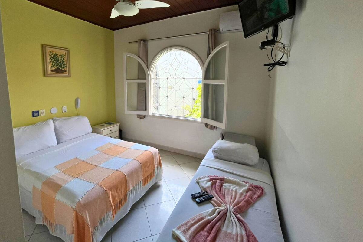 Une chambre d’hôtel comprenant un lit, un lit plus petit ou un canapé, une fenêtre avec des volets décoratifs, un ventilateur de plafond, un téléviseur fixé au mur et une table de chevet avec une lampe.